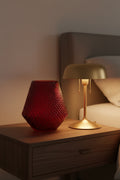 Deep crimson hobnail glass vase on a bedside table beside a lit brass lamp