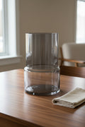 Plain smoked grey glass vase on a walnut dining table beside a folded linen napkin