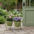 Sage green bench plant stand holding two flowering pots on a stone patio beside a garden door