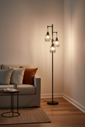 Three-light floor lamp lit in a living room corner beside a linen sofa